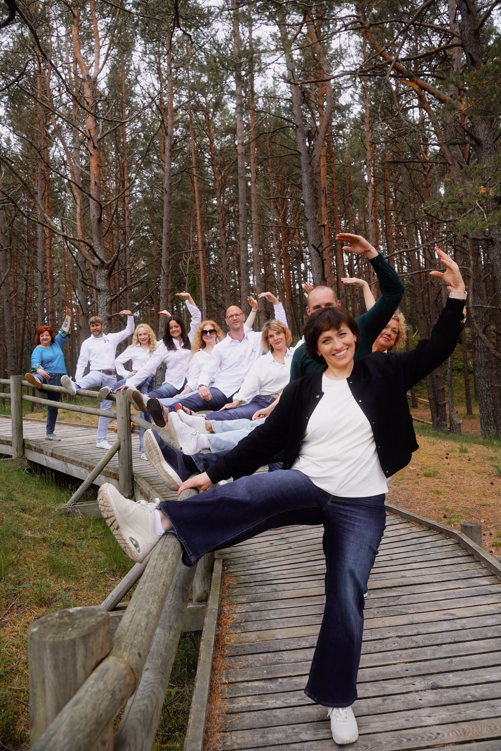 Desmit cilvēku grupa meža laukumā pozē, ceļot rokas un kājas jautrā pozā uz koka tiltiņa.