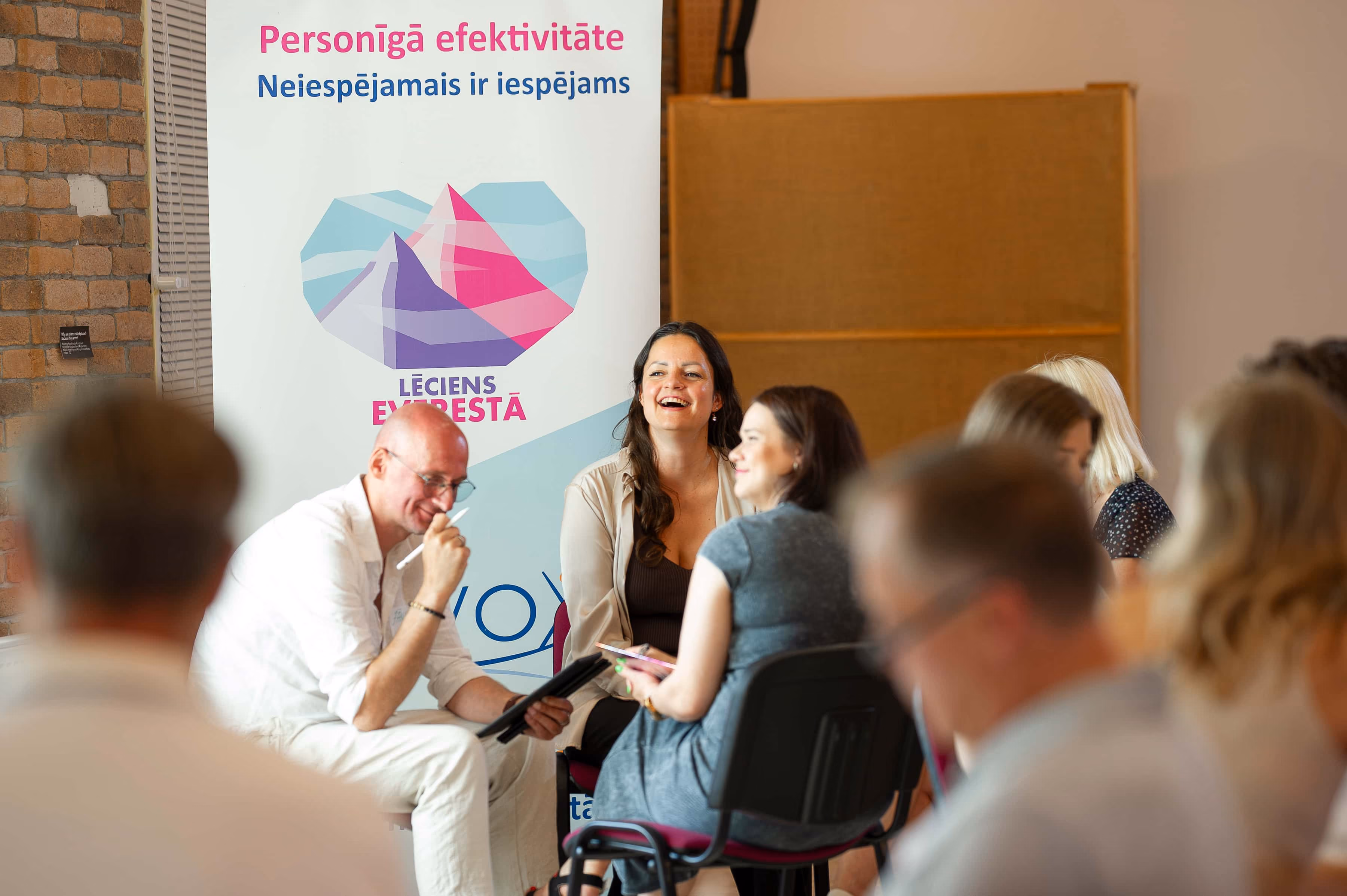 Cilvēki sarunājas un smejas seminārā, fonā plakāts ar uzrakstu Personīgā efektivitāte.