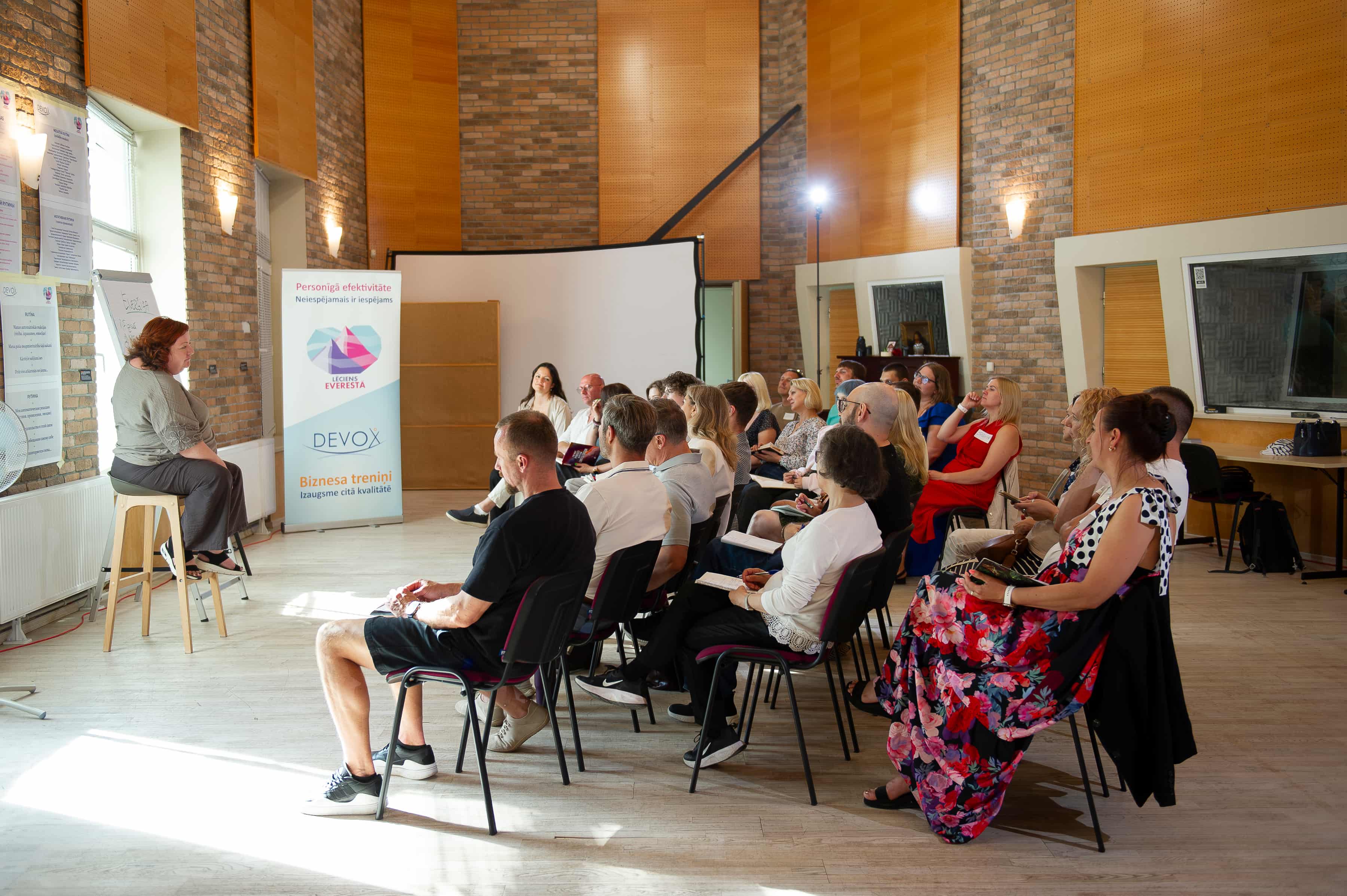 Lekcija ar sievieti uz krēsla runā auditorijai, kas klausās seminārā ar plakātu un projektoru.