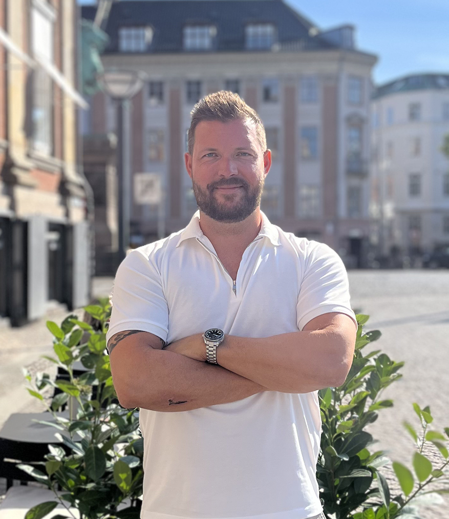Man with beard in white polo shirt standing with arms crossed outdoors with buildings and plants behind him.