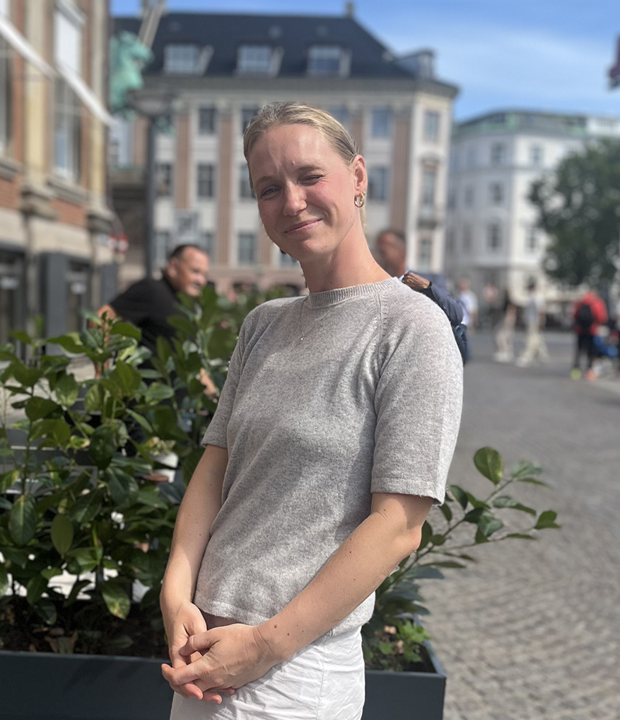 Woman in a gray shirt and white pants standing outside on a sunny day with buildings behind her.
