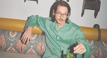 Man with glasses and mustache wearing green, holding a drink while sitting on a patterned couch.