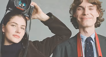 Two young adults smiling, one holding a camera above her head, the other with a camera strap.