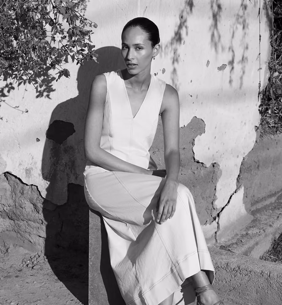 Woman sitting on a block against a cracked wall, wearing a sleeveless dress and sandals.