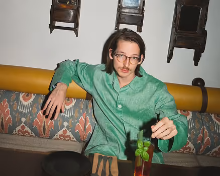 Man in green shirt sitting on patterned bench, holding a glass with a green garnish and black straw.