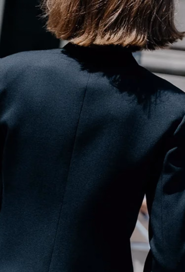 Person with short brown hair wearing a black textured garment, seen from the back in an outdoor setting.