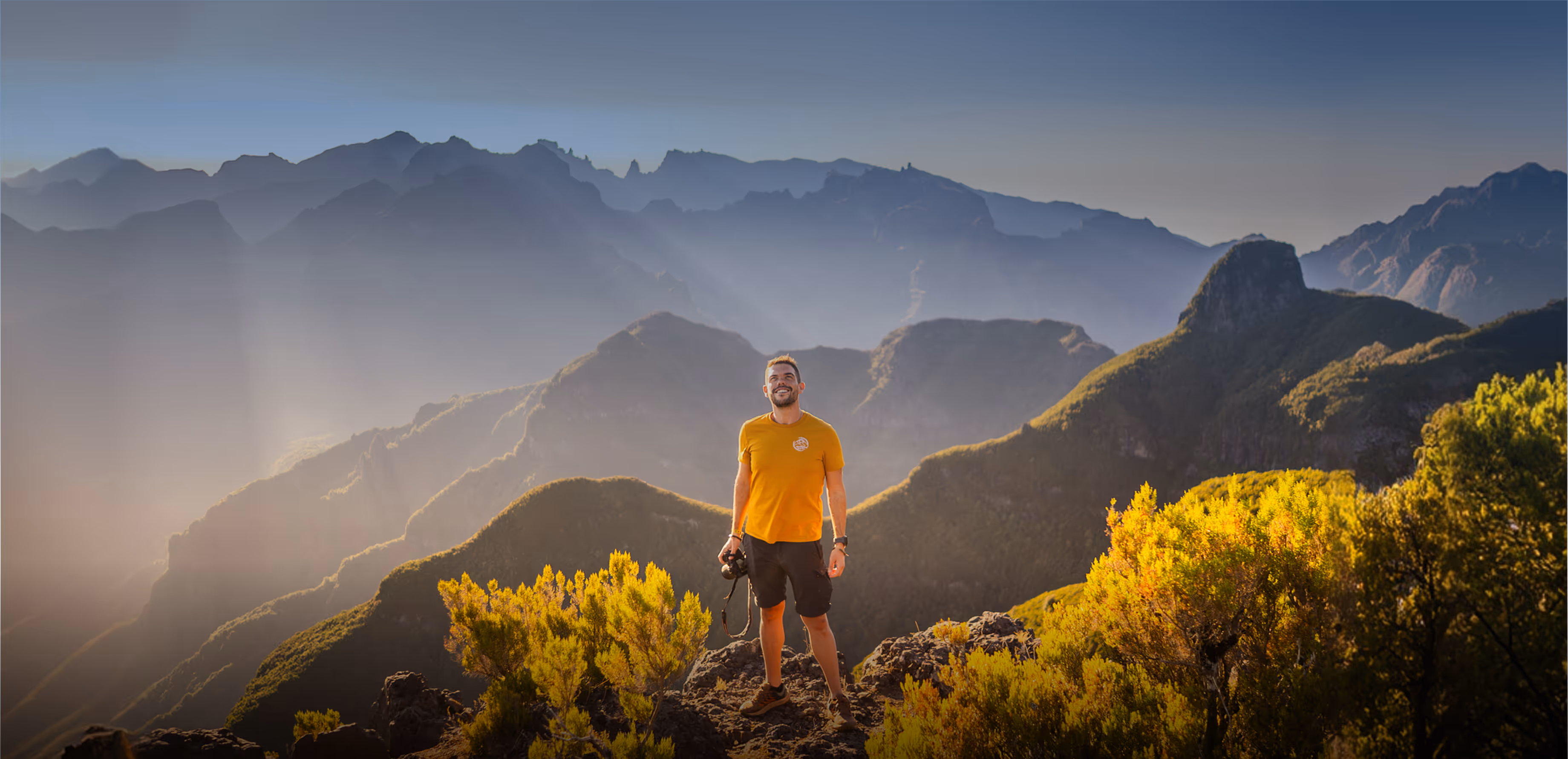 Man in orange shirt standing on rocky mountain peak amid yellow-green shrubs and distant misty mountains.