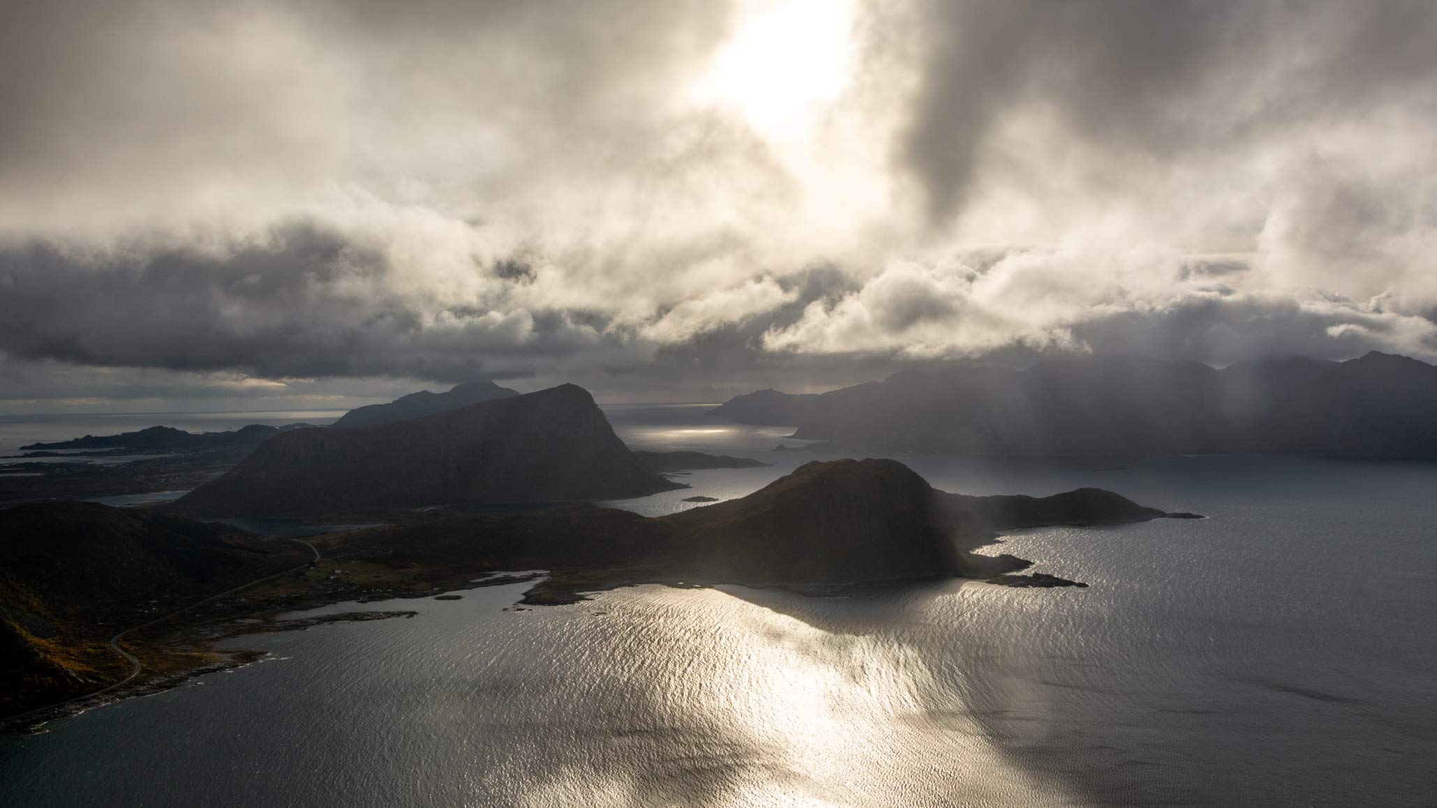 Norsk kyst med fjell og hav under dramatisk skydekke med sollys som skinner gjennom skyene.