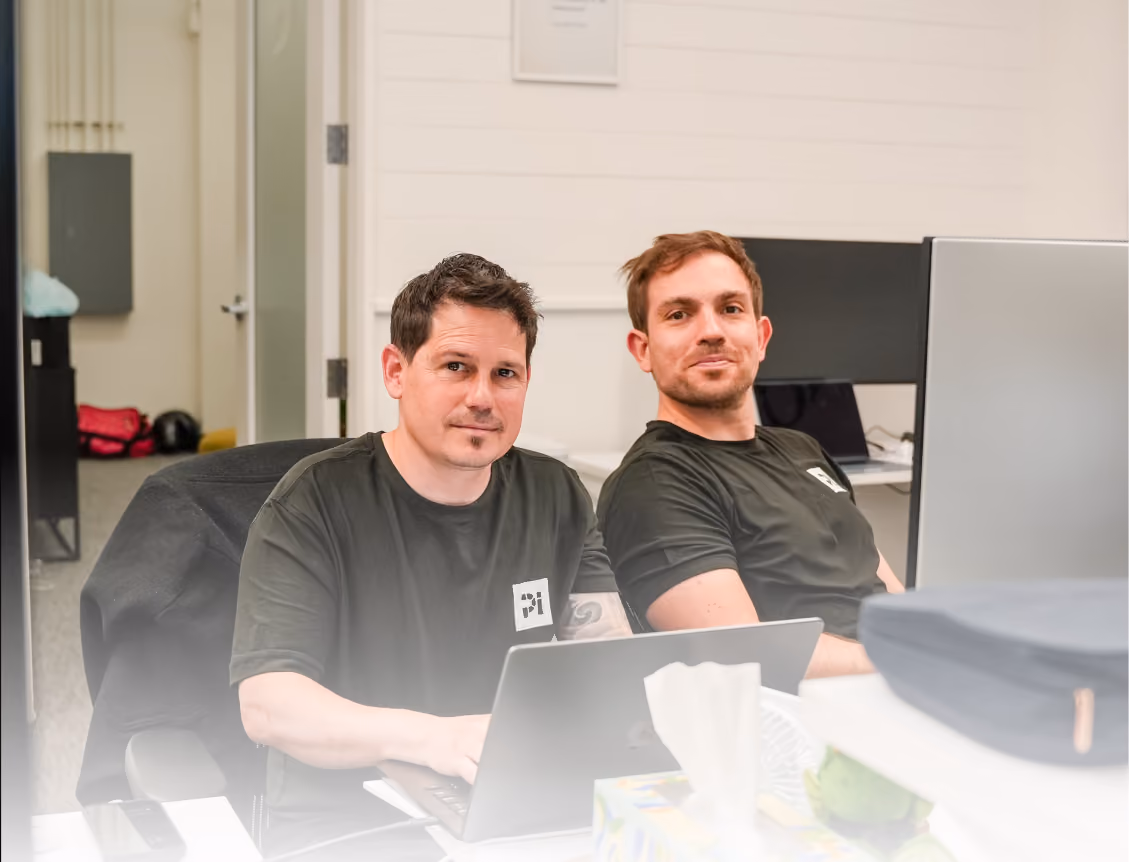 Two men sitting at a desk with laptops in an office, both wearing black t-shirts and looking at the camera.