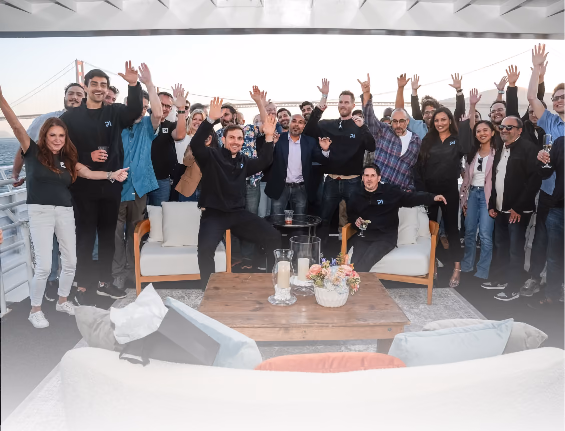 Group of people smiling and raising hands on a boat near a suspension bridge at sunset.