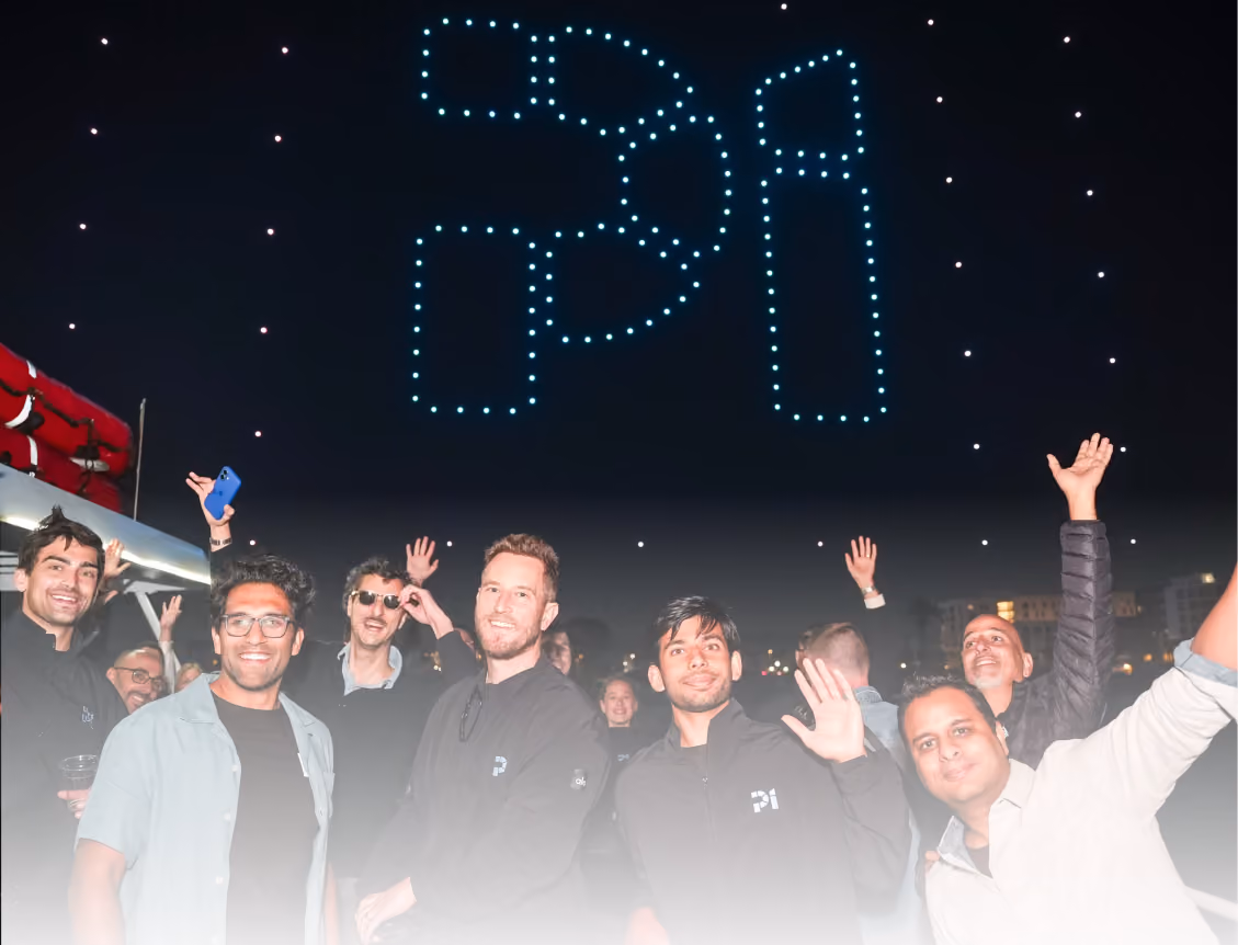 Group of people smiling and waving at night under drones forming the letters 'pi' in the sky.