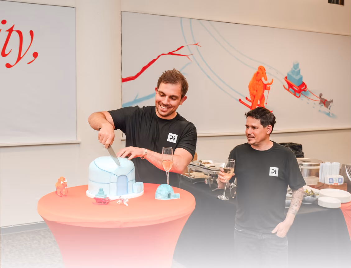 Man cutting igloo-shaped cake while another man holds a drink, both wearing black shirts indoors.