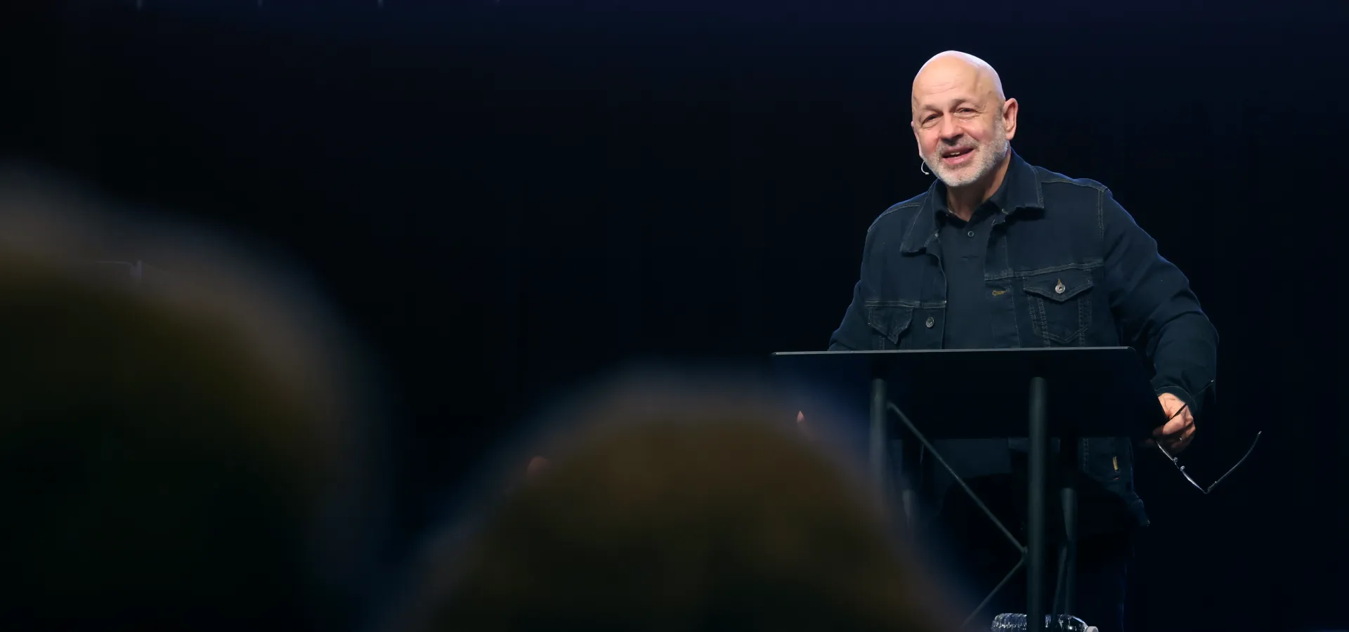 Smiling elderly man speaking on stage behind a podium with dark background.