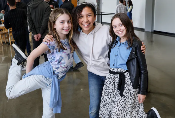 Three smiling girls posing indoors, two with one leg lifted, wearing casual and stylish clothes.