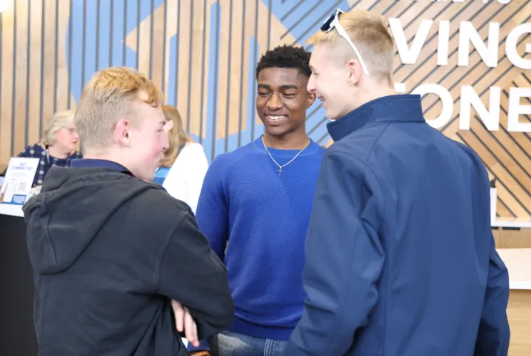 Three teenage boys smiling and talking indoors near a wooden wall with partially visible text.