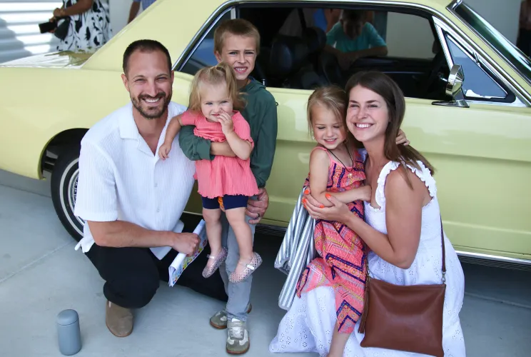 Smiling family of five with two kids posing next to a yellow vintage car.