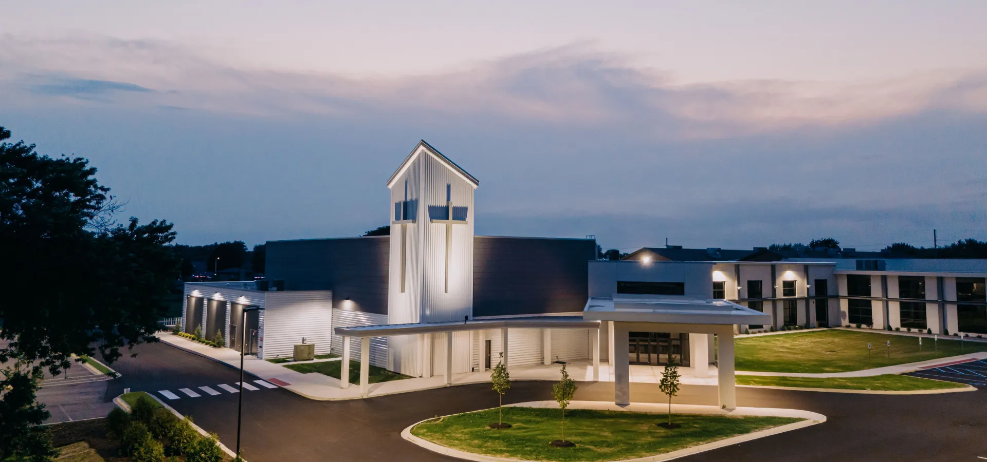 Modern church building at dusk with lit crosses on a tall white tower and surrounding greenery.