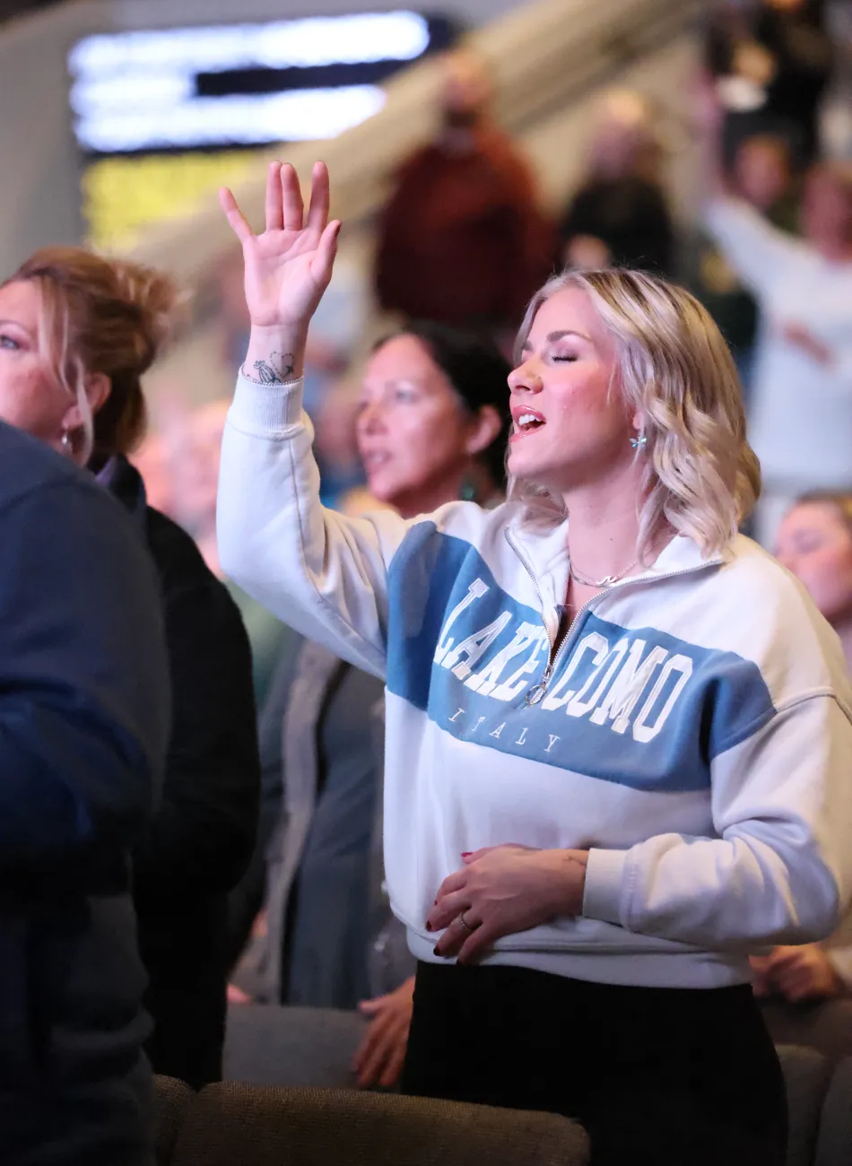 Woman with blond hair raising her hand and singing with eyes closed at a gathering.
