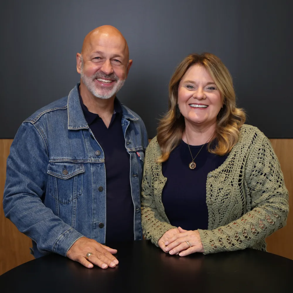 Smiling man in a denim jacket and woman in a green knit cardigan posing at a black table.