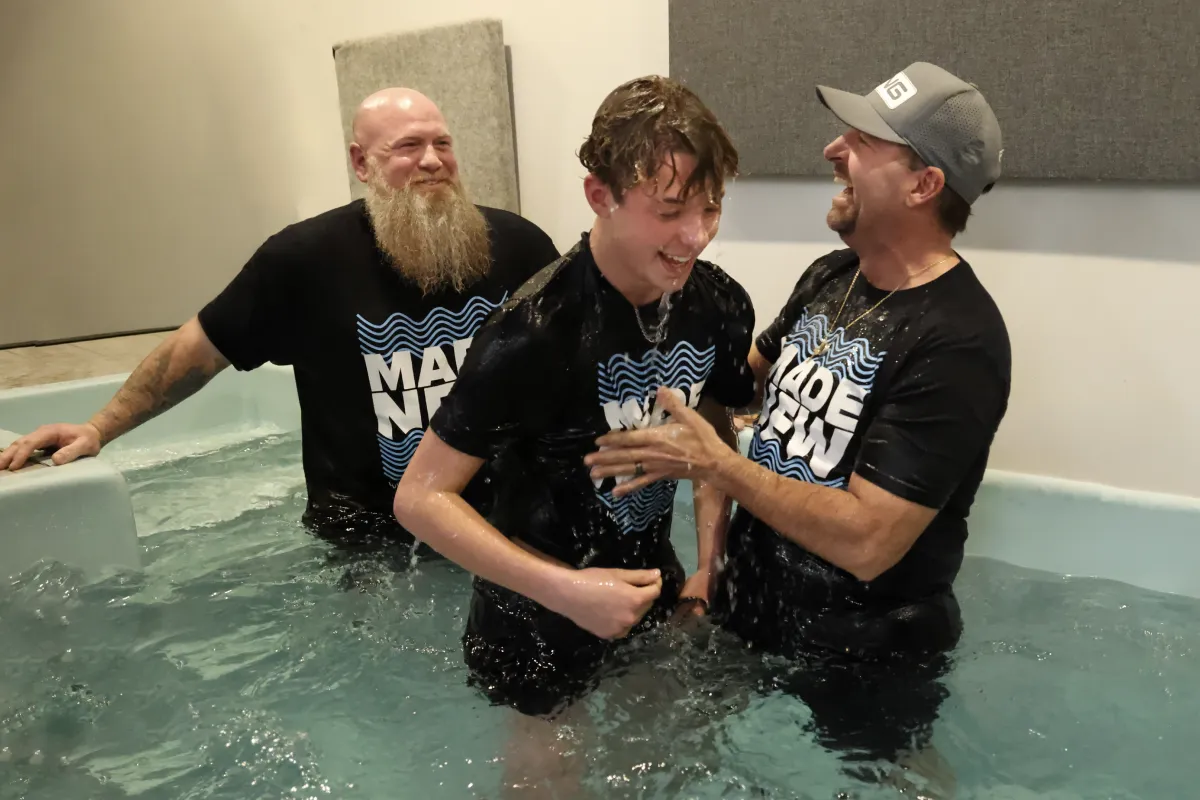 A boy smiling in a baptismal pool with two men in matching black 'Made New' shirts.