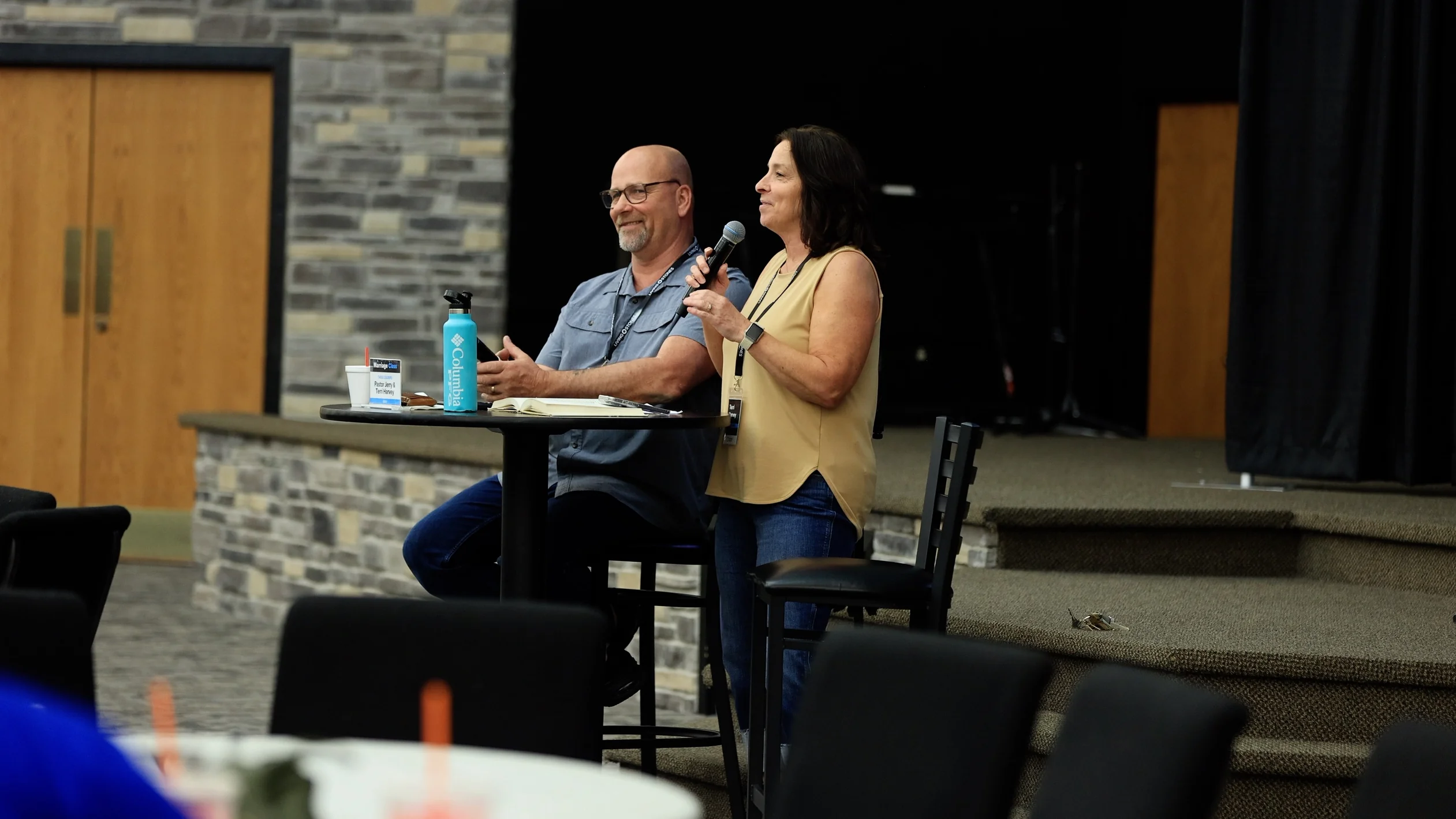 Man and woman presenting at an event, woman speaking into a microphone, man seated at table.