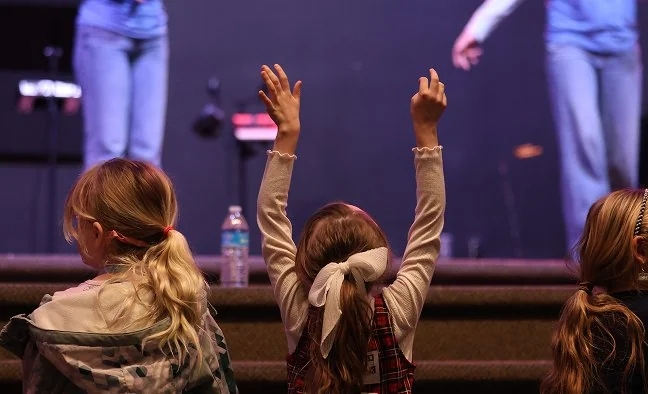 Children with raised hands at a worship event, focusing on the back of a girl with a white bow in her hair.
