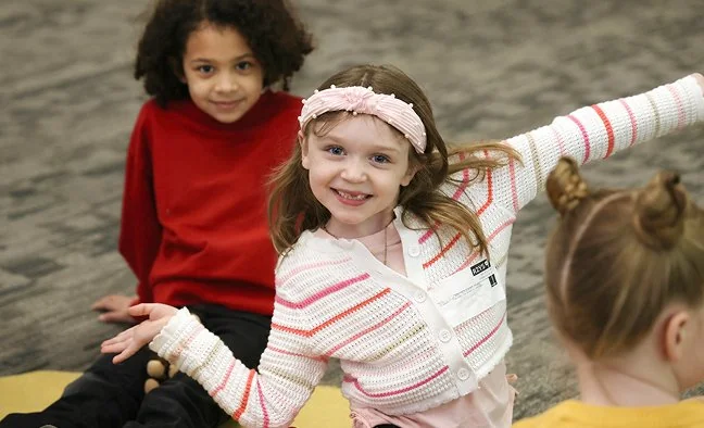 Three children indoors; one girl in white striped sweater smiling with arms outstretched.