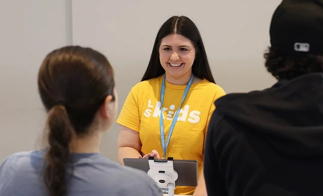 Woman in a yellow shirt with 'kids' lanyard smiles while checking in two volunteers at a tablet station.