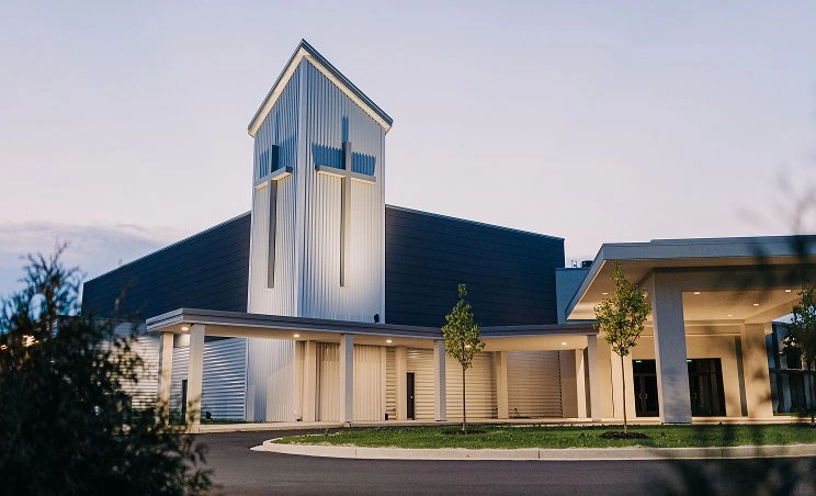 Modern church building with illuminated cross tower at dusk and small trees in front.