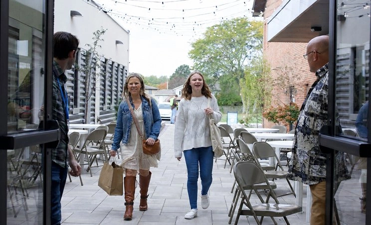 Two women walking on outdoor patio with string lights, greeted by two men by open doors.