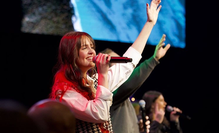 Young woman singing with microphone and raising her hand on stage during worship.