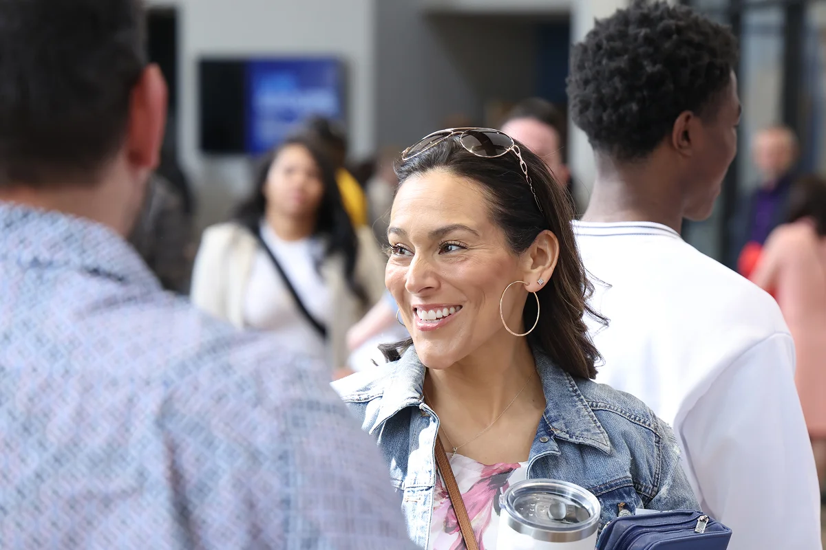 Smiling woman in denim jacket holding a travel mug and purse, talking to a man in blurred crowd.