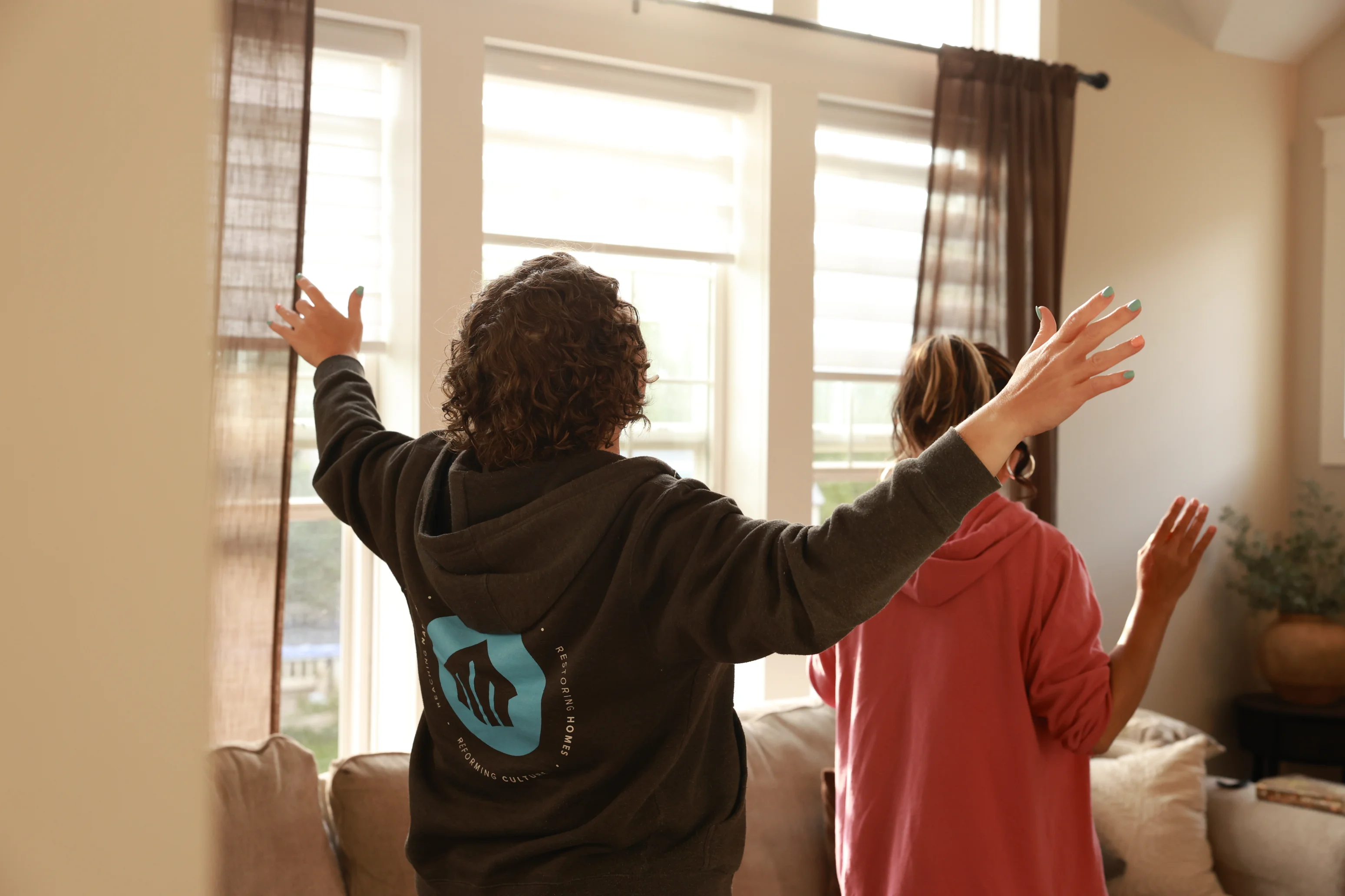 Two people with raised arms facing sunlit windows in a cozy living room with a couch and curtains.