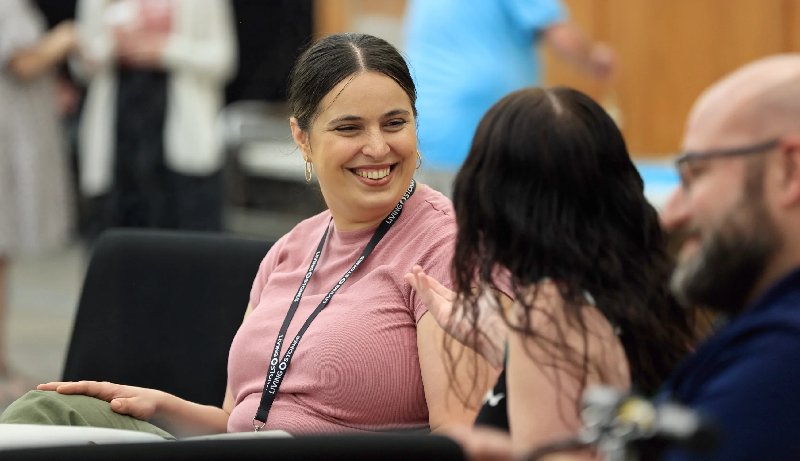 Smiling woman wearing a pink shirt and lanyard talks with two people in a casual indoor setting.