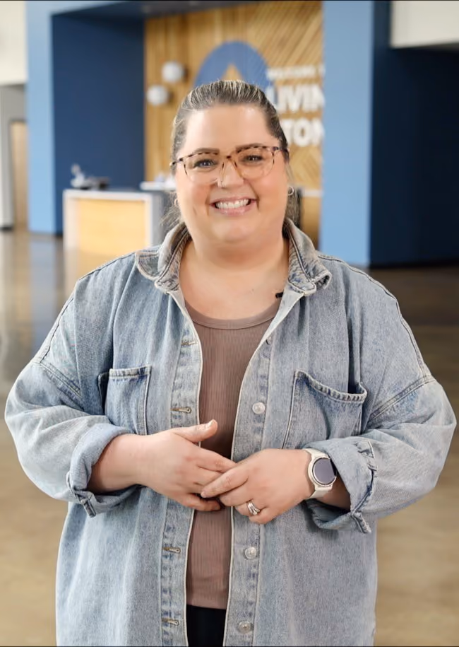 Smiling woman with glasses wearing a denim jacket and smartwatch standing indoors.