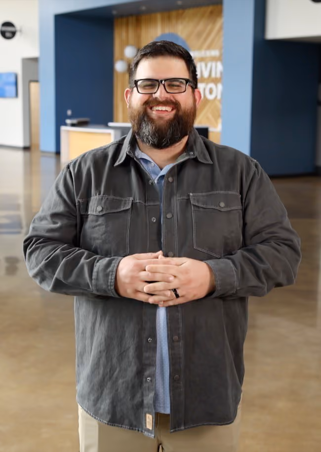 Smiling man with glasses and beard, wearing a gray jacket, standing indoors with hands clasped.