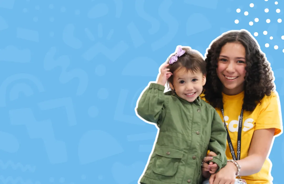 Smiling young woman in yellow with curly hair holds happy toddler in green jacket on blue background.