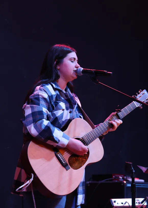 Woman in a plaid shirt singing and playing acoustic guitar on stage at a Living Stones youth event.