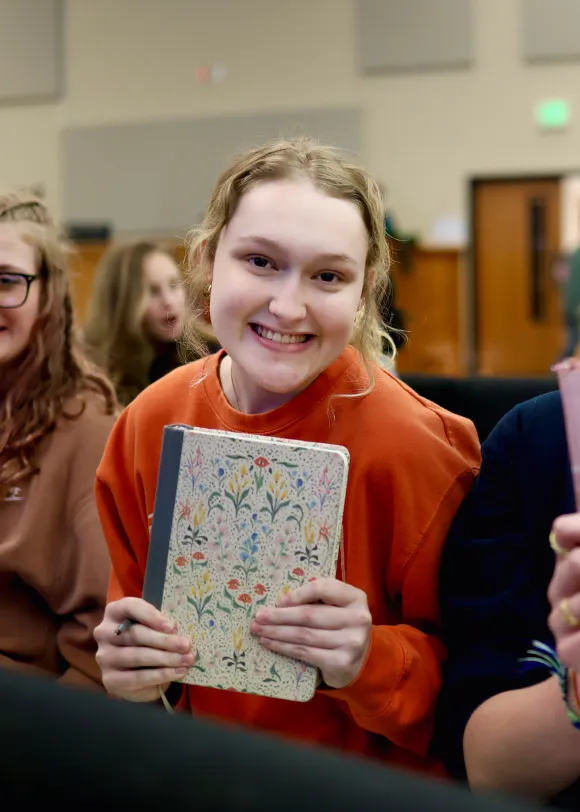 Smiling young woman in an orange sweater holding a floral notebook at a Living Stones youth event.
