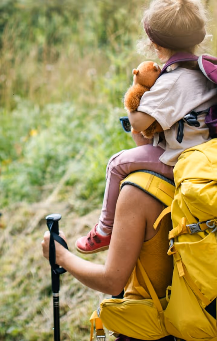 Erwachsener trägt Kind mit Teddybär auf den Schultern beim Wandern mit Stock und großem Rucksack.