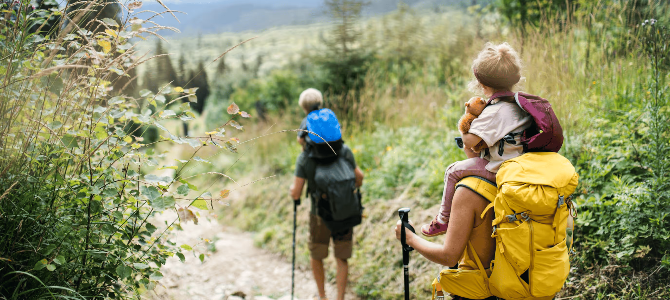 Wanderer mit gelbem Rucksack trägt Kind, das Kuscheltier hält, auf schmalem Waldpfad.