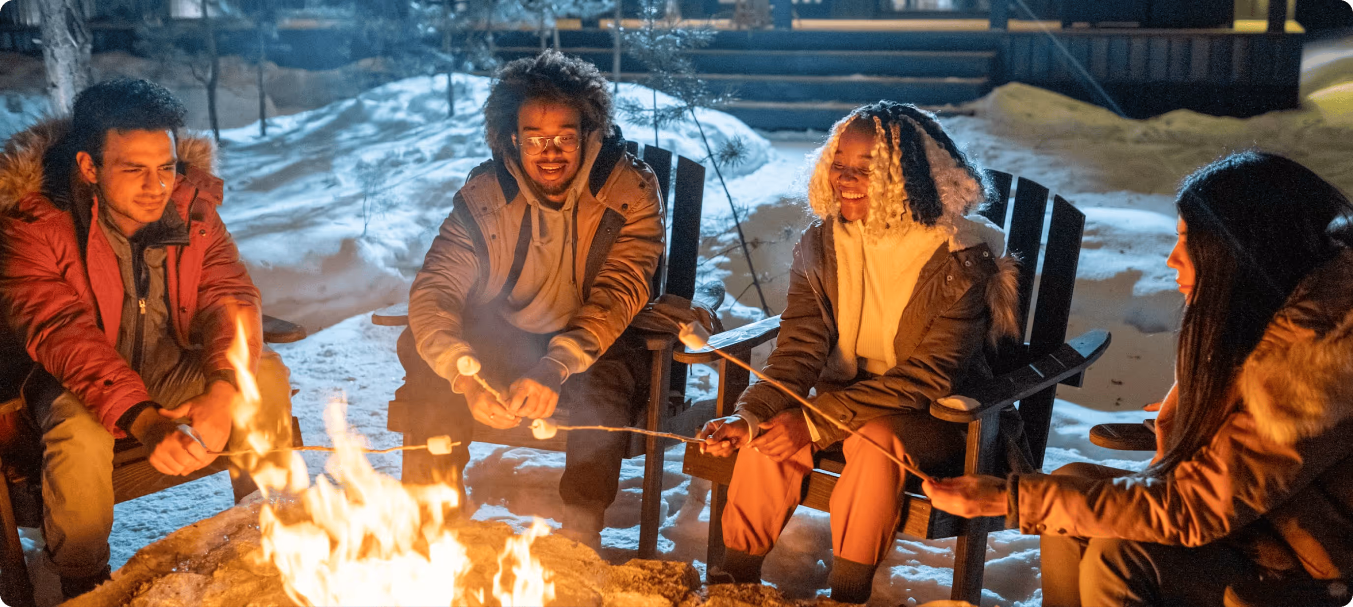Vier Freunde rösten Marshmallows am Lagerfeuer in einer verschneiten Winterlandschaft.
