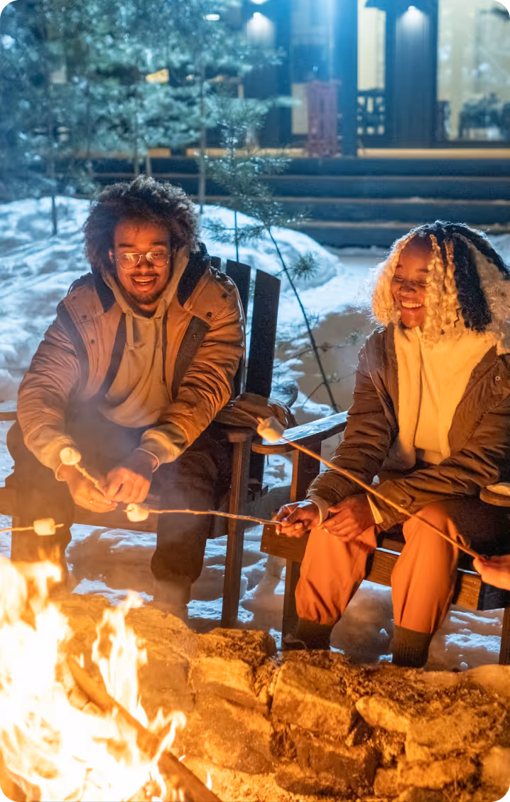 Zwei lächelnde Personen rösten Marshmallows über einem Lagerfeuer im Schnee bei Nacht.