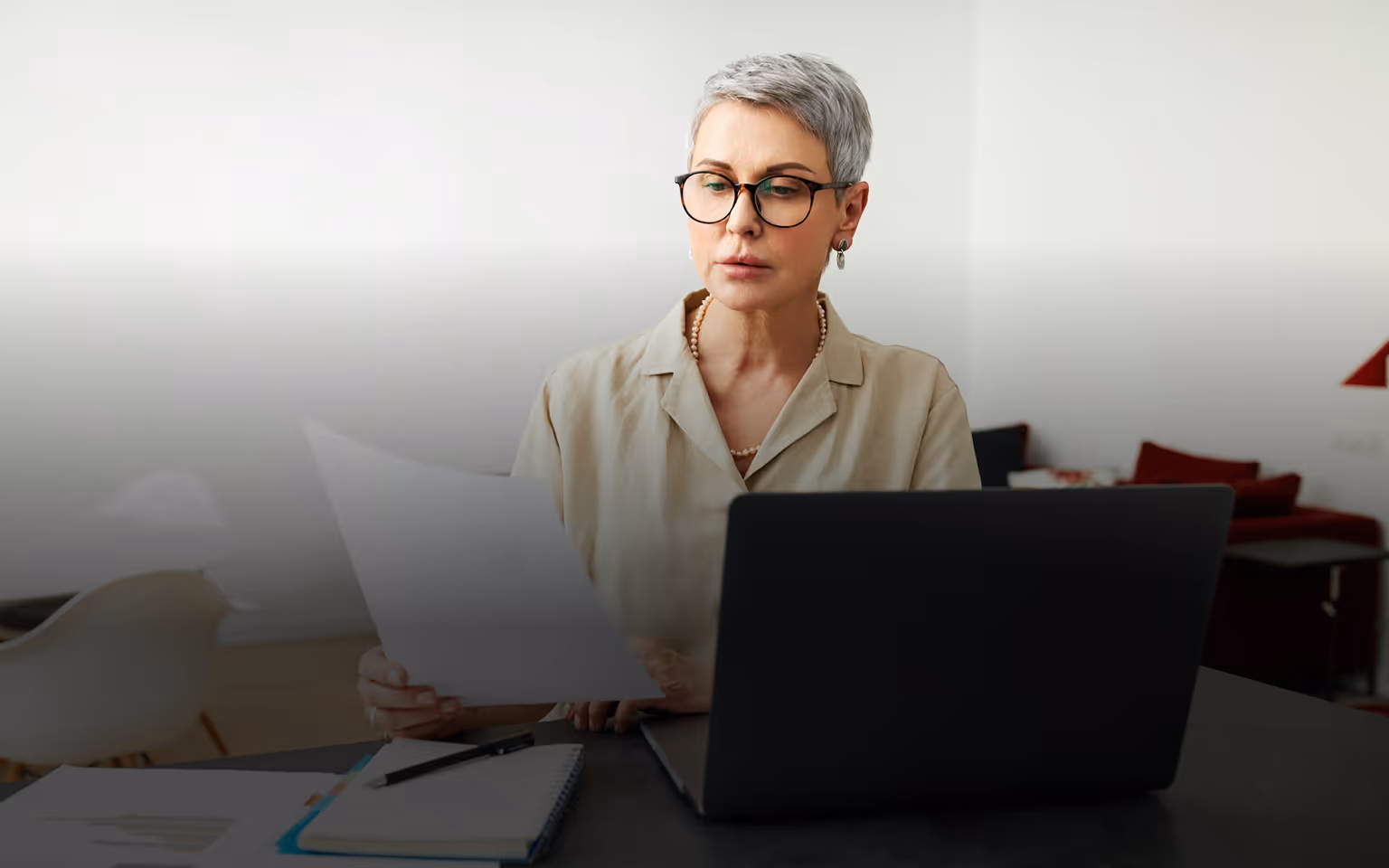 Frau mit kurzen grauen Haaren und Brille liest ein Dokument am Laptop in modernem Büro.