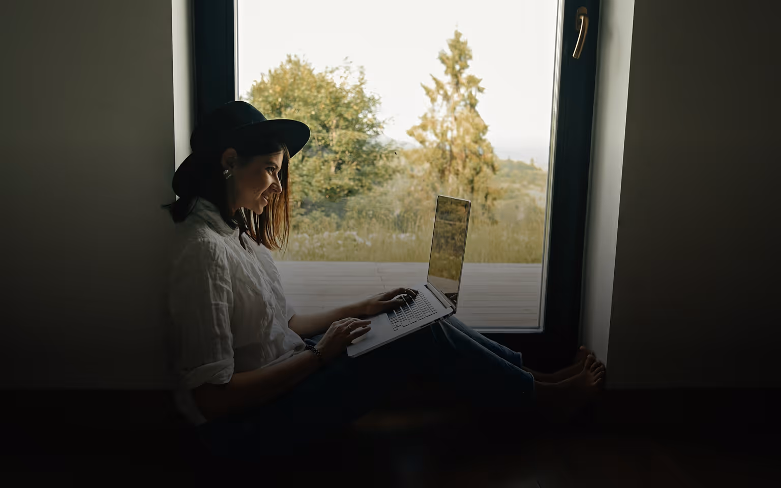 Frau mit Hut sitzt am Fenster und arbeitet an einem Laptop mit Blick nach draußen.