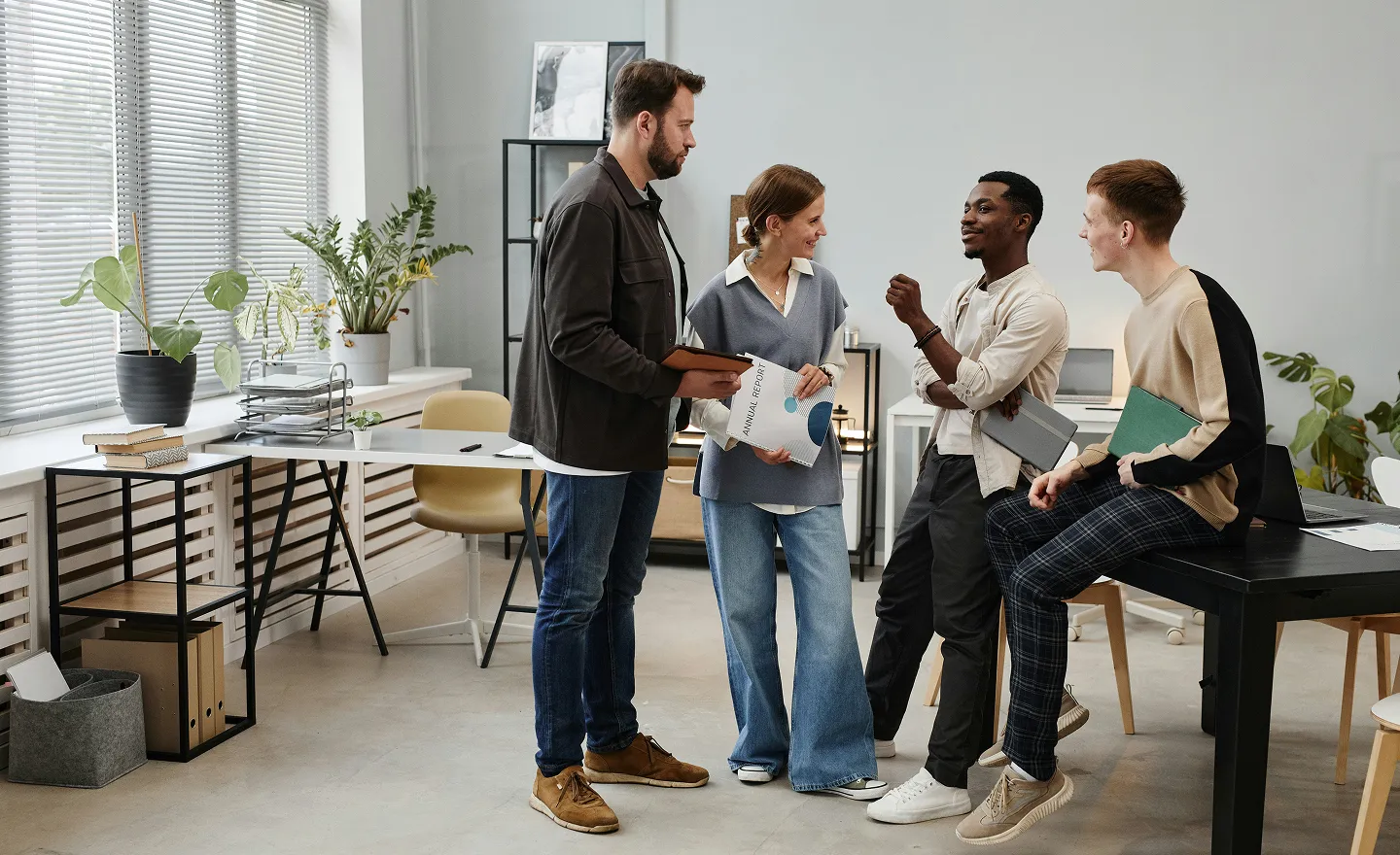 Vier junge Erwachsene diskutieren lächelnd in einem modernen Büro mit Pflanzen und Schreibtischen.