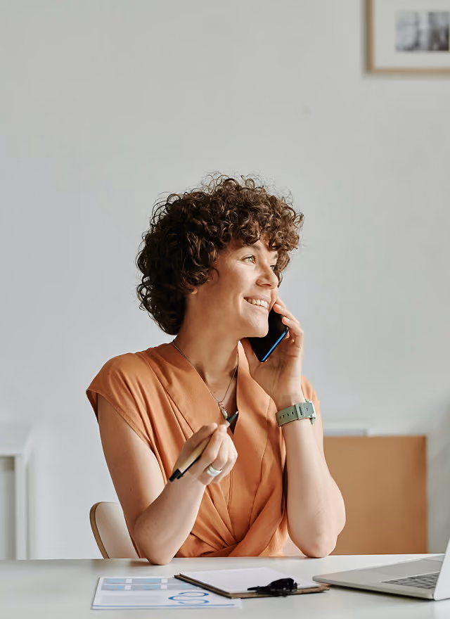 Frau mit lockigem Haar spricht lächelnd am Telefon an einem Schreibtisch mit Laptop und Unterlagen.