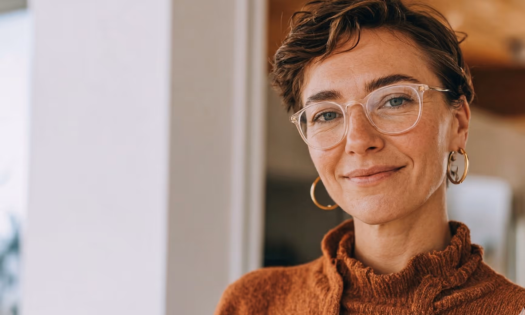 Femme souriante aux cheveux courts, lunettes transparentes et pull marron à col montant.