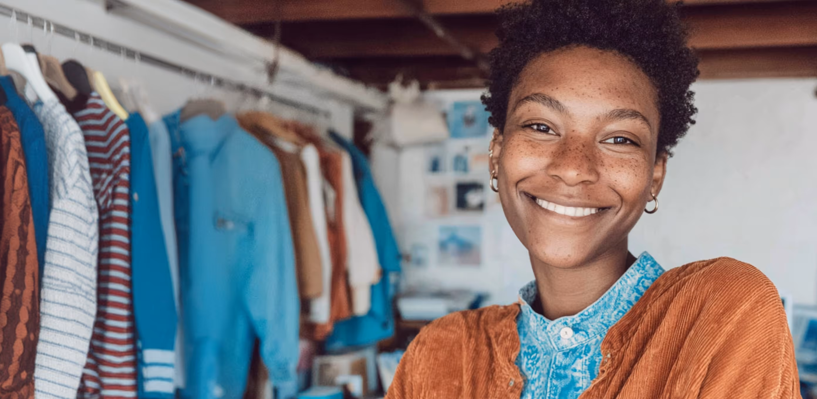 Jeune femme souriante avec cheveux bouclés et taches de rousseur devant un portant de vêtements colorés.