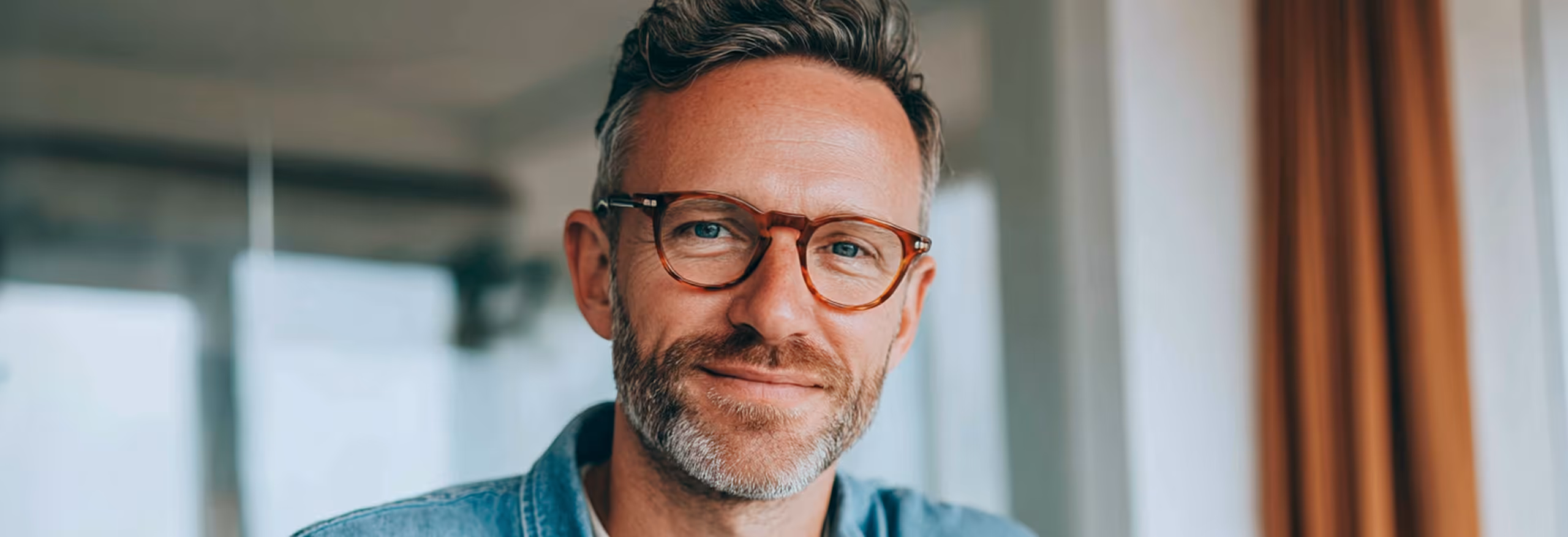 Homme souriant avec des lunettes et une barbe grise, à l'intérieur près d'une fenêtre.